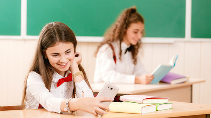 two schoolgirls are sitting at the desk in the classroom using a telephone