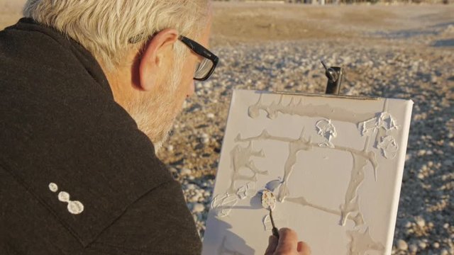 Senior man paints a picture on the beach. Back view of elderly male artist applying paint to canvas with a spatula at pebble sunrise sea beach.