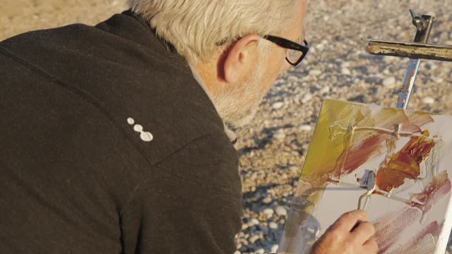 Senior man paints a picture on the beach. Over the shoulder shot of elderly male artist applying paint to canvas with a spatula at pebble sunrise sea beach.