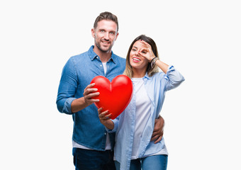 Young couple in love holding red heart over isolated background with happy face smiling doing ok sign with hand on eye looking through fingers