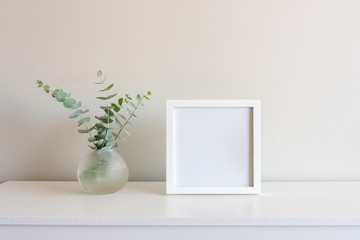 Eucalyptus leaves in small round glass vase next to blank square picture frame on white shelf...