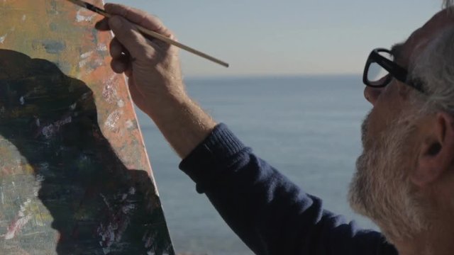 Senior man paints a picture on the beach. Elderly male artist painting modern abstract picture with brush on morning calm sea background. Close-up.