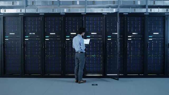  In the Modern Data Center: IT Engineer Standing Beside Open Server Rack Cabinets, Does Wireless Maintenance and Diagnostics Procedure with a Laptop. Zoom Out Camera Shot.
