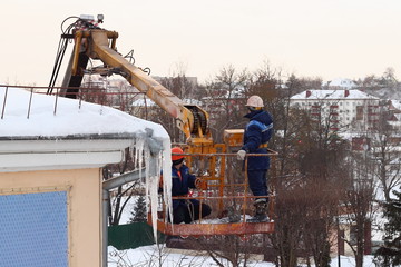 Workers in overalls and helmets on the crane basket remove icicles from roof of the house on a...