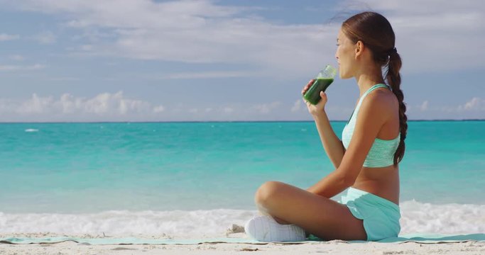 Fitness woman drinking a healthy green smoothie or cold pressed juice for a detox weight loss cleanse. Happy sporty asian girl on beach taking a snack break with a vegan beverage for a vegetarian diet