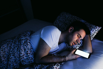 Young man using smartphone in bed at night - obrazy, fototapety, plakaty