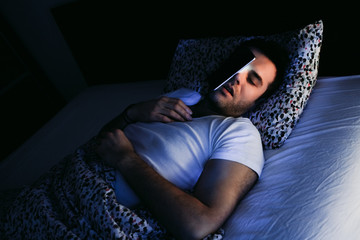 Young man using smartphone in bed at night - obrazy, fototapety, plakaty