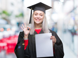 Young blonde woman wearing graduate uniform holding degree over isolated background doing ok sign...