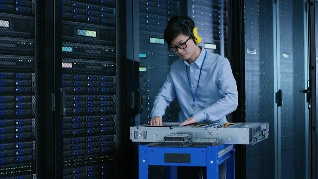 In Data Center: IT Engineer Wearing Protective Muffs Installs New Hardware for Server Rack. Specialist Doing Maintenance, Running Diagnostics and Updating Hardware for Stable Functioning of a System.