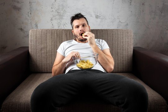 The guy in the shirt is lying on the couch, eating chips and watching a sports channel. The concept of laziness, frustration, procrastination, the person at home.