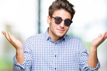 Young handsome man wearing sunglasses over isolated background clueless and confused expression...