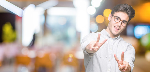 Plakat Young handsome man wearing glasses over isolated background smiling looking to the camera showing fingers doing victory sign. Number two.