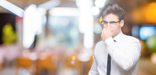 Young business man wearing glasses over isolated background smelling something stinky and disgusting, intolerable smell, holding breath with fingers on nose. Bad smells concept.