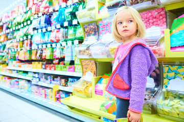 Adorable child blond girl select sweets on shelves in supermarket 