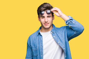 Young handsome man wearing sunglasses over isolated background winking looking at the camera with sexy expression, cheerful and happy face.