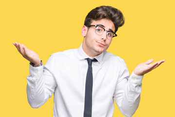 Young business man wearing glasses over isolated background clueless and confused expression with arms and hands raised. Doubt concept.