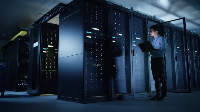 In Data Center: Male IT Technician Running Maintenance Programme on a Laptop, Controls Operational Server Rack Optimal Functioning. Modern High-Tech Telecommunications Operational Super Computer.