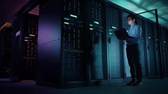 In Data Center: Male IT Technician Running Maintenance Programme on a Laptop, Controls Operational Server Rack Optimal Functioning. Modern High-Tech Telecommunications Operational Super Computer.