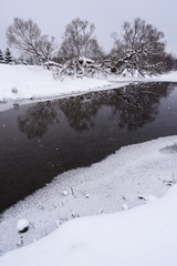 Dull sunless winter landscape. Dark river in ice and trees on snowy banks