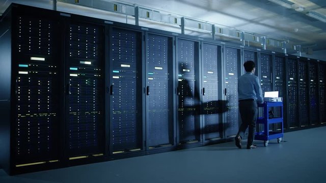  In the Modern Data Center: IT Engineer Doing Maintenance and Diagnostics. Pushes Cart alongside Server Racks, Opens Cabinet to Install New Hardware for Planned System Update, Equipment Replacement.