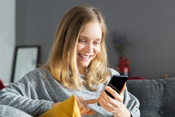 Cheerful phone user excited with new mobile app. Positive young woman using smartphone in living room. Mobile app concept