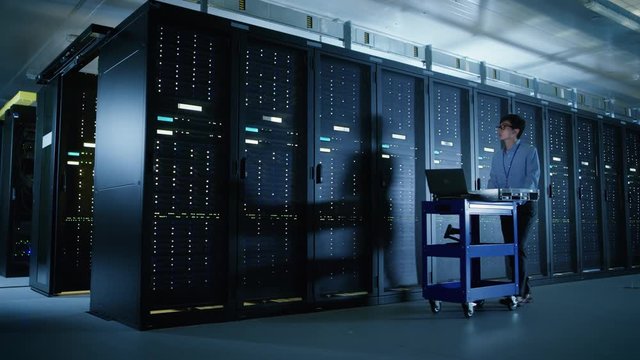  In the Modern Data Center: IT Engineer Doing Maintenance and Diagnostics. Pushes Cart alongside Server Racks, Ready to Install New Hardware for Planned System Update, Equipment Replacement.