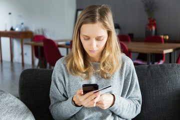 Focused customer paying online. Serious young woman sitting on sofa at home, using smartphone and holding credit card. Online payment concept