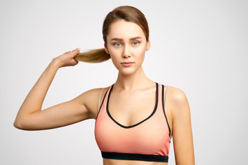 A sporty young woman with a serious face and a confident expression looks at the camera and squeezes in her hand a bundle of her long hair. Isolated Studio portrait on white background