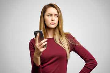 Naklejka premium Gloomy upset girl makes grimace from despair, holds smartphone, reads disapponting news, looks dejected. Isolated on white background in Studio