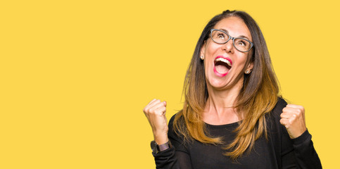 Beautiful middle age woman wearing glasses very happy and excited doing winner gesture with arms...