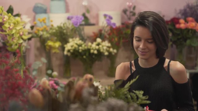 Very beautiful girl with short black hair, hairstyle bob, in black shoulderless sweater admiring and touching bouquet/roses/nature/chamomiles/cloves/lilies in flower shop. Smelling floral atmosphere
