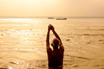 evocative view of old man performing daily puja ritual on calm water of ganges river at sunrise seen from behind, with warm light - obrazy, fototapety, plakaty