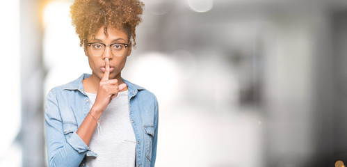 Beautiful young african american woman wearing glasses over isolated background asking to be quiet with finger on lips. Silence and secret concept.