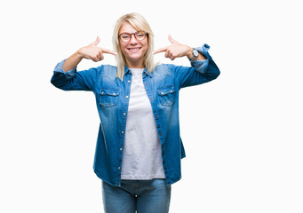 Obraz premium Young beautiful blonde woman wearing glasses over isolated background smiling confident showing and pointing with fingers teeth and mouth. Health concept.