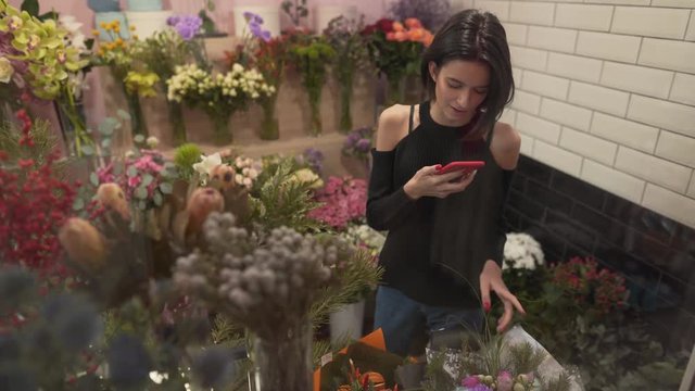 Very beautiful brunette girl with short black hair, hairstyle bob, in black shoulderless sweater and jeans is in flower shop, touching with her gentle hands bouquet of flowers, taking picture of them