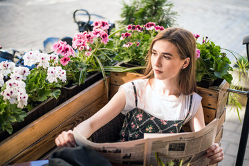 Young woman having a breakfast reading newspaper outdoors at the typical french cafe terrace in France. Playful girl with long hair posing outdoor. Wearing wool cap. Street fashion look. Cute teen