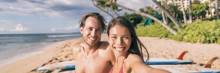 Fototapeta premium Beach vacation selfie couple. Happy young interracial friends taking photo with phone after surfing class on tropical Hawaii beach. Banner panorama. Travel vacation destination watersports.