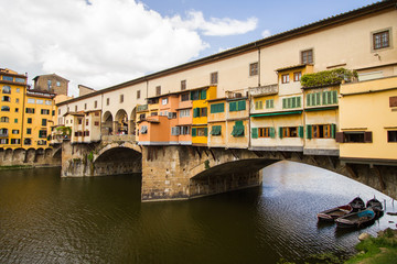 Fototapeta na wymiar The Shopping Bridge with Boats Moored - Boats await their customers under colorful Ponte Vecchio - a bridge hosting many jewelry shops - that crosses the River Arno. Florence, Italy