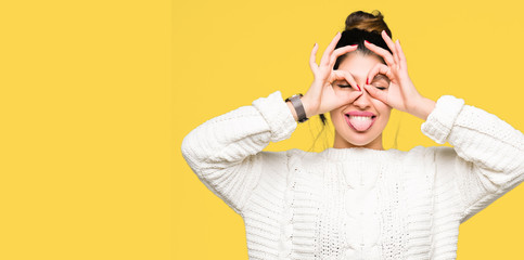 Young beautiful woman wearing winter sweater doing ok gesture like binoculars sticking tongue out, eyes looking through fingers. Crazy expression.