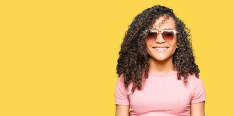 Young beautiful woman with curly hair wearing pink sunglasses with a happy and cool smile on face. Lucky person.