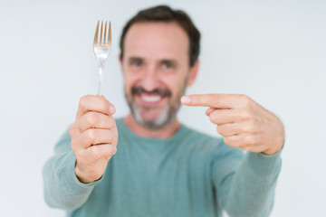 Senior man holding silver fork over isolated background very happy pointing with hand and finger