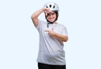 Young adult cyclist woman with down syndrome wearing safety helmet over isolated background smiling making frame with hands and fingers with happy face. Creativity and photography concept.