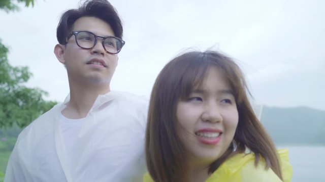 Slow motion - Young Asian sweet couple feeling happy using romantic time playing rain while wearing raincoat standing near lake. Lifestyle couple enjoy and relax in rainy day.