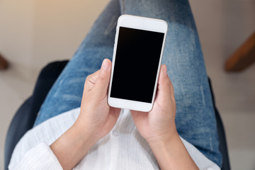 Top view mockup image of hands holding and using a white mobile phone with blank screen while sitting