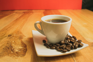 cafe en tasa blanca con granos de cafe sobre un plato blanco, en una mesa de madera con un fondo de pared roja  