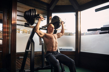 one man, 30-39 years, bodybuilder exercising overhead plate press machine. At home (with many windows) in his own gym with professional fitness equipment.