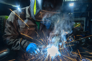 Man welder in welding mask, building uniform and blue protective gloves welds metal car muffler...