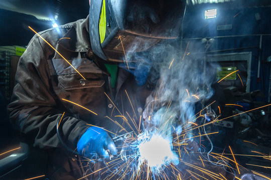 Man Welder In Welding Mask, Building Uniform And Blue Protective Gloves Welds Metal Car Muffler With Welding Machine In Auto Repair Shop, In The Background Construction Site