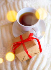 A cup with tea or coffee and a box with a gift tied with a red ribbon on a white knitted background. Romantic breakfast in bed for Valentine's Day or a gift for mother's day
