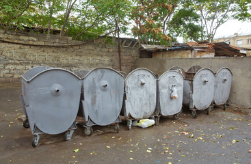 A row of gray iron garbage cans on the street. Grey garbage containers in the City on the asphalt on the background of red building - Image